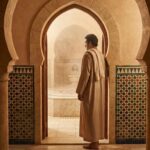 Seorang pria berpakaian jubah tradisional Muslim berdiri di ambang pintu pemandian umum (hammam) beruap dengan arsitektur Islam yang khas, mencerminkan adab dan refleksi sebelum masuk