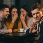 Ilustrasi seorang pria/wanita yang menunjukkan ekspresi tidak nyaman dan memberi isyarat tangan menolak saat teman-temannya sedang asyik tertawa menggunjing orang lain.