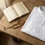 Baju koko putih bersih terlipat rapi di samping kitab doa Al-Qur'an terbuka dan tasbih kayu di atas meja kayu dengan cahaya matahari pagi.