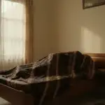 Suasana hening di kamar tidur dengan jenazah yang sudah ditutup kain batik panjang dari kepala hingga kaki, ilustrasi persiapan awal merawat jenazah sebelum dimandikan.