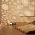 Foto bernuansa hangat yang menampilkan peralatan terapi bekam (hijamah) berupa gelas kop dan pompa di atas meja kayu, di samping buku antik terbuka bertuliskan aksara Arab. Di latar belakang, terdapat kalender dinding yang melingkari tanggal Hijriah 17, 19, dan 21 dengan warna merah, serta mangkuk berisi herbal kering.