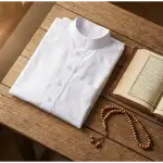 Foto flat lay baju koko putih terlipat rapi di samping tasbih kayu dan buku kitab terbuka di atas meja kayu, berlatar cahaya alami. Ilustrasi adab berpakaian Islami.