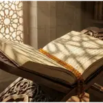 An open classical Islamic manuscript resting on a carved wooden rihal (book stand) with prayer beads, illuminated by sunlight through a patterned window.