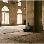 A Muslim man sitting silently in a grand mosque, demonstrating the proper etiquette of I'tikaf through quiet contemplation and reverence.