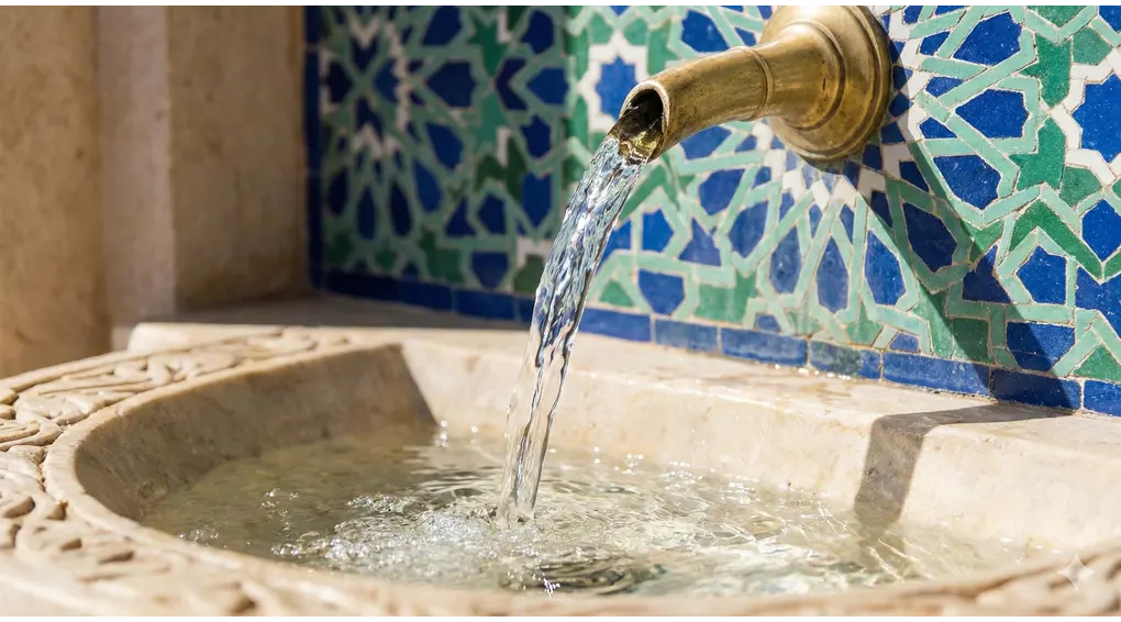 The Mandatory Pillars of Ghusl: 2 Obligatory Conditions in Shafi'i Fiqh 3 Clear, pure water flowing vigorously from a brass spout into a stone basin against a background of blue and green Islamic mosaic tiles.