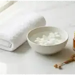 A serene flat-lay photograph on marble showing items symbolic of Islamic purification: a clean white towel, a bowl of water with flowers, a small bottle of musk perfume oil, and tasbih prayer beads.