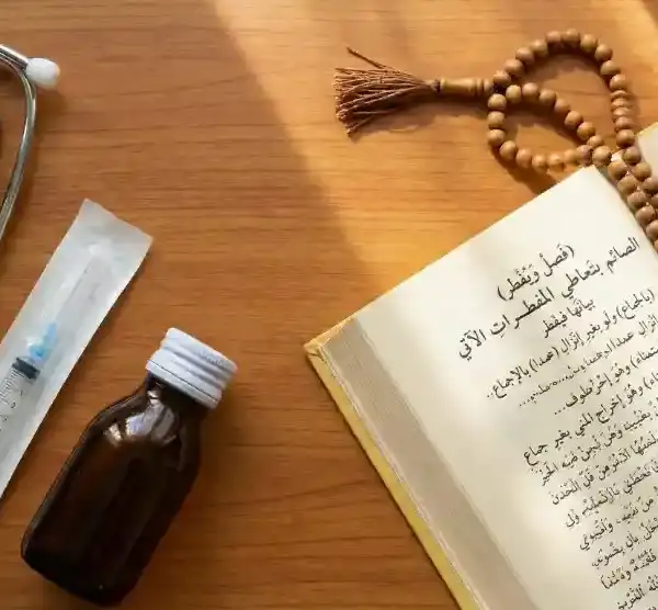 Peralatan medis seperti stetoskop, suntikan, dan obat botol di samping kitab kuning terbuka dengan tasbih di atas meja kayu.