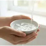 Tangan seseorang memegang mangkuk keramik berisi air jernih dengan kelopak bunga putih mengapung, di bawah aliran air kran yang lembut dan cahaya jendela yang terang.