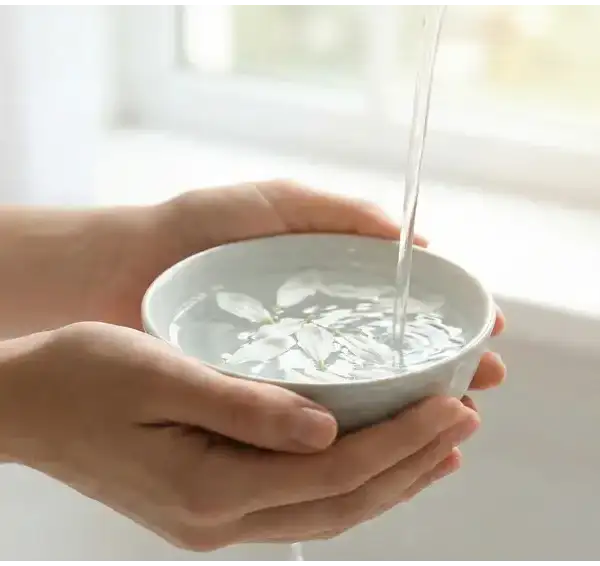 Tangan seseorang memegang mangkuk keramik berisi air jernih dengan kelopak bunga putih mengapung, di bawah aliran air kran yang lembut dan cahaya jendela yang terang.