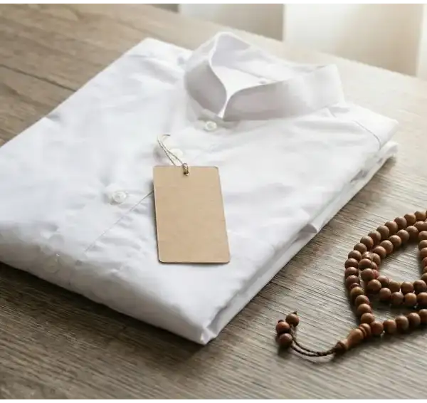 Baju kemeja putih baru terlipat rapi dengan label tag kosong dan tasbih kayu di atas meja kayu, ilustrasi doa alhamdulillahilladhi kasani hadha