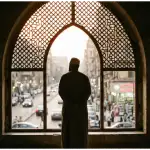 A contemplative photograph from inside a dimly lit mosque showing the silhouette of a Muslim man standing near an arched window looking outside, symbolizing the spiritual boundary of I'tikaf.
