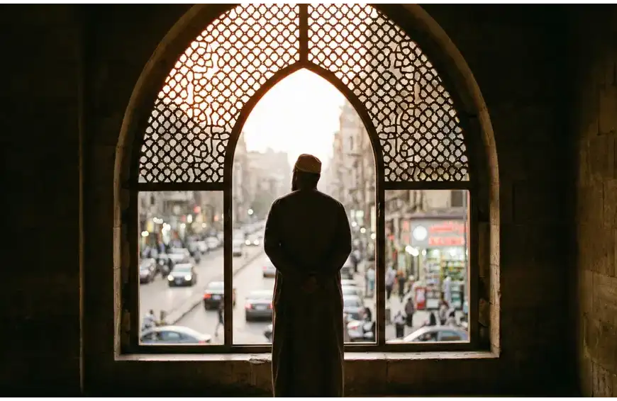 Home 31 A contemplative photograph from inside a dimly lit mosque showing the silhouette of a Muslim man standing near an arched window looking outside, symbolizing the spiritual boundary of I'tikaf.