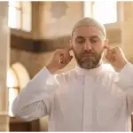 Adhan and Iqamah Recitation Guide: Rules & Translation (Shafi'i Fiqh) A medium close-up photo of a Muslim muadhin in a mosque, calling the Adhan with a focused expression and placing his index fingers in his ears according to the Sunnah.