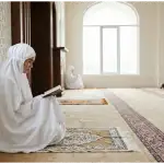 Foto seorang wanita muslimah duduk khusyuk membaca Al-Qur'an di sudut area khusus wanita di dalam masjid yang tenang dengan pencahayaan alami, menggambarkan suasana i'tikaf.