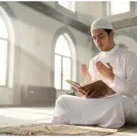 A student of knowledge dressed in a clean white koko shirt sitting with proper etiquette in a brightly lit mosque prior to a majlis of knowledge.