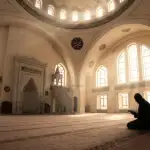 Foto sudut lebar interior masjid megah dengan cahaya matahari yang menembus masuk melalui kubah dan jendela, memperlihatkan siluet seorang pria Muslim duduk khusyuk membaca Al-Qur'an dalam suasana hening i'tikaf.