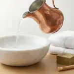 A traditional copper jug pouring clear, fresh water into a white marble basin, accompanied by clean white towels, olive soap, and a miswak stick on a wooden surface, symbolizing preparation for Islamic ritual purification.
