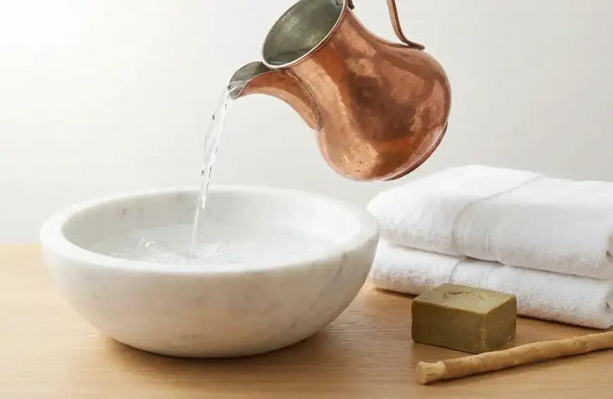Home 7 A traditional copper jug pouring clear, fresh water into a white marble basin, accompanied by clean white towels, olive soap, and a miswak stick on a wooden surface, symbolizing preparation for Islamic ritual purification.