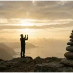 Silhouette of a person praying with hands raised toward a golden sunrise on a coastal hill, next to a balanced stone cairn symbolizing a strong spiritual foundation.