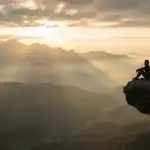 A silhouette of a man sitting on a cliff edge at sunrise, overlooking vast misty mountains, representing deep spiritual reflection or tafakur in Islam.