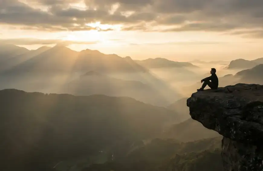 Home 7 A silhouette of a man sitting on a cliff edge at sunrise, overlooking vast misty mountains, representing deep spiritual reflection or tafakur in Islam.