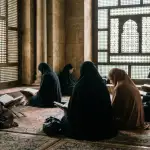 A documentary-style photograph of Muslim women in hijabs quietly reading the Quran and praying behind wooden mashrabiya screens in the dedicated women's section of a historic mosque.