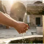Close-up of a person's hands performing wudu (ablution) with clear flowing water at an ancient mosque stone basin during a golden sunset.