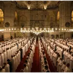 A massive congregation of Muslims standing in orderly rows performing the Tarawih prayer inside a grand, beautifully illuminated mosque during the month of Ramadan.