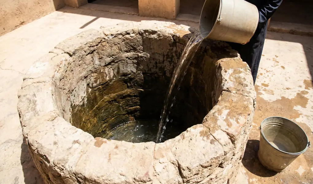 How to Handle and Purifying Well Water Contaminated by Najis: Insights from Asna al-Matalib 2 Clean water is being poured heavily from a metal bucket into a stone well whose water level appears low (less than two qullahs). This illustrates the at-taktsir technique for purifying small amounts of impure water.