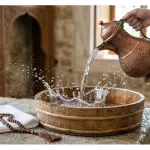 Crystal-clear water being poured from a classic copper pitcher into a wooden basin for thaharah preparation.