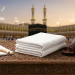 Two unstitched white ihram cloths, a pair of open sandals, and a miswak on a carved wooden table with the silhouette of Masjid al-Haram in the background.