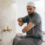 A Muslim man pouring water from a copper vessel onto his arm to perform wudu in a modern mosque purification area with gold Arabic calligraphy.